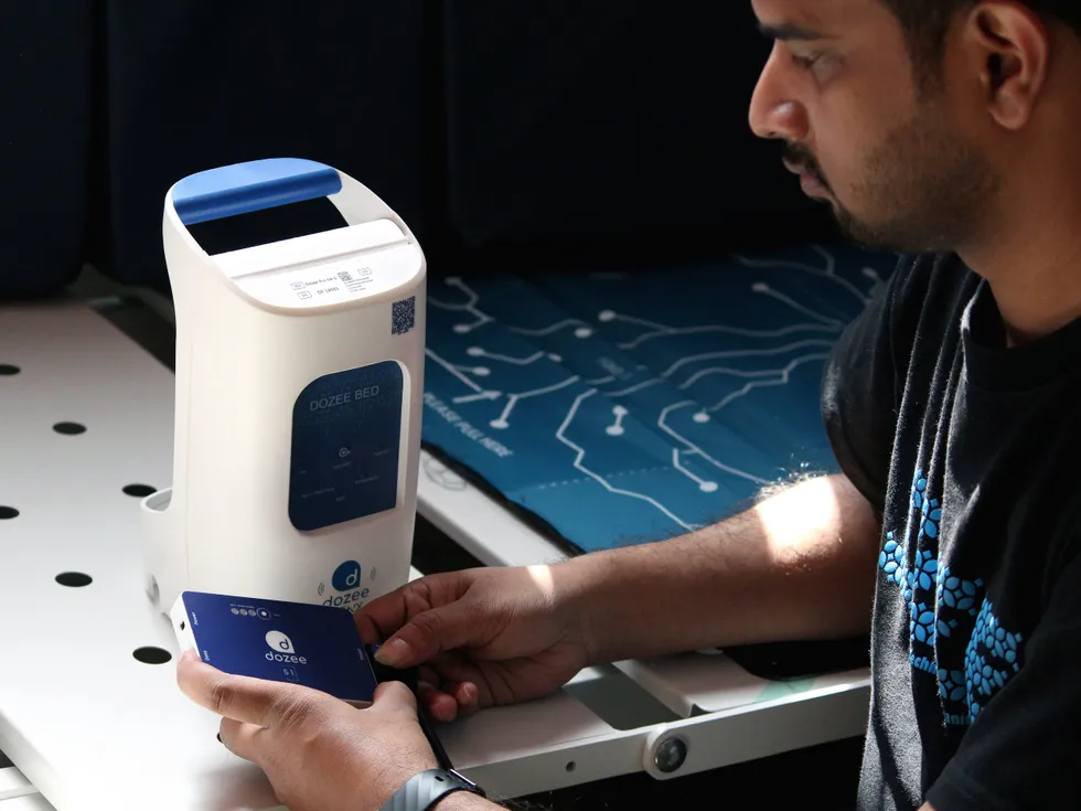An adult Indian man looking at a rectangular device in his hand, labeled \u201cdozee\u201d.