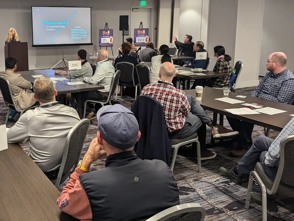 A woman standing next to a presentation screen while speaking to small seated groups during a professional workshop.