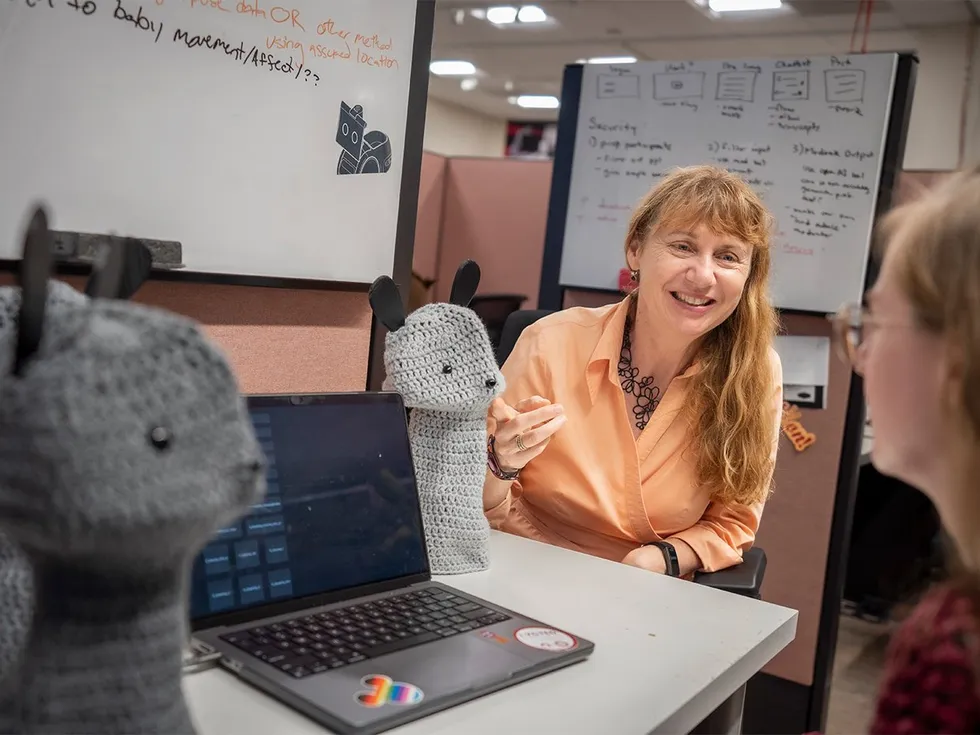 A smiling blonde woman gestures at a customizable tabletop robot that wears a knit outfit of a cute animal over its shell.