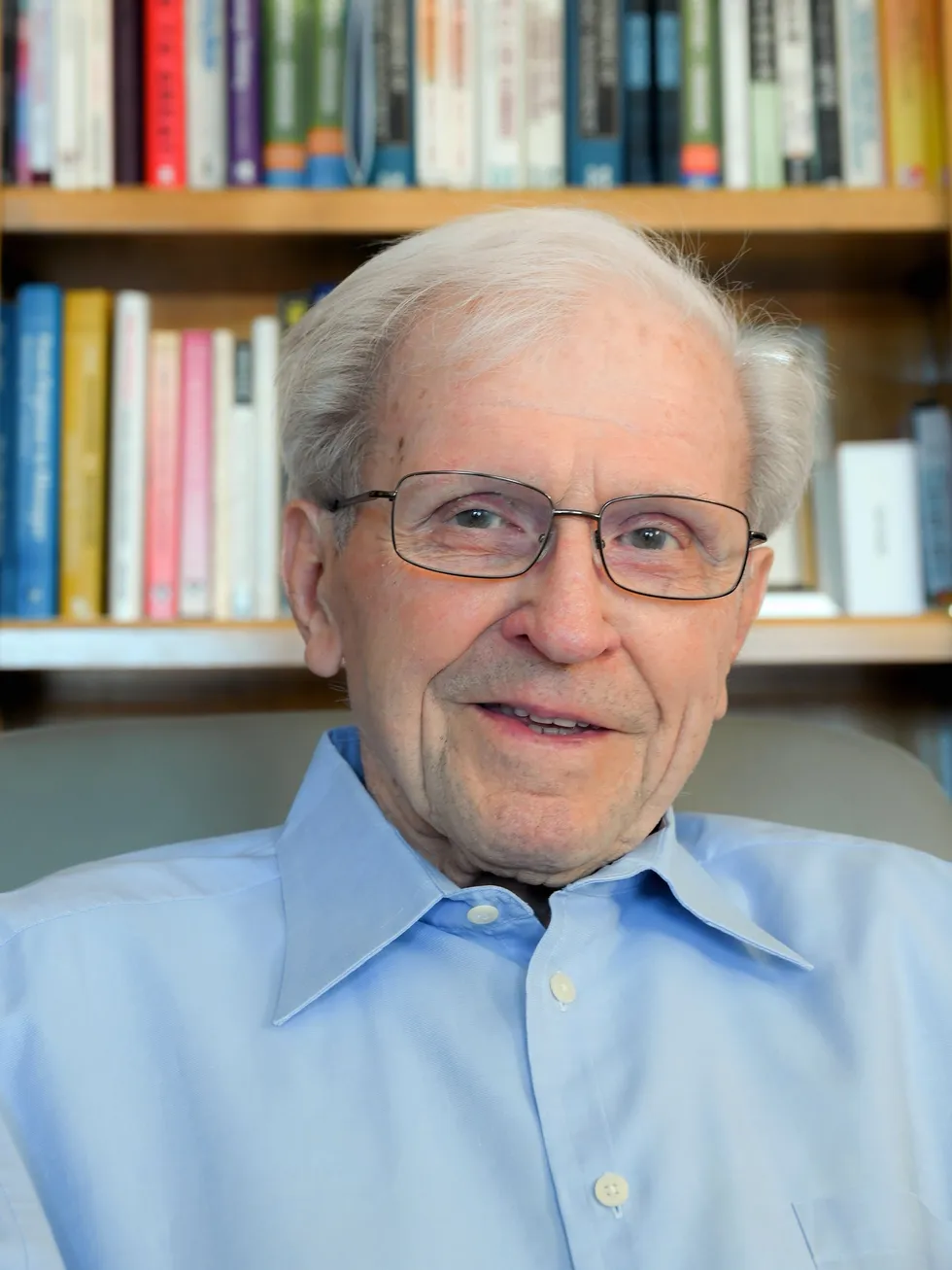 An elderly white man smiling in a dress shirt against a background of bookshelves.